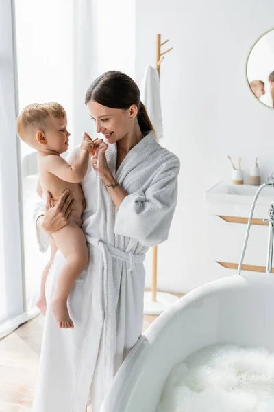 Joyeuse mère en peignoir tenant dans les bras et les doigts touchants avec tout-petit fils près de la baignoire avec mousse de bain — Photo de stock