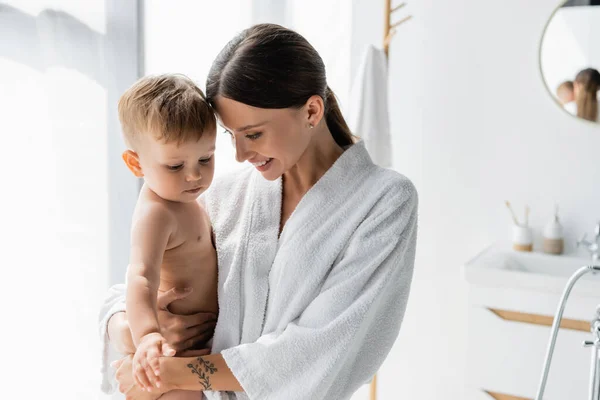 Cheerful and tattooed mother in bathrobe holding in arms toddler son — Stock Photo