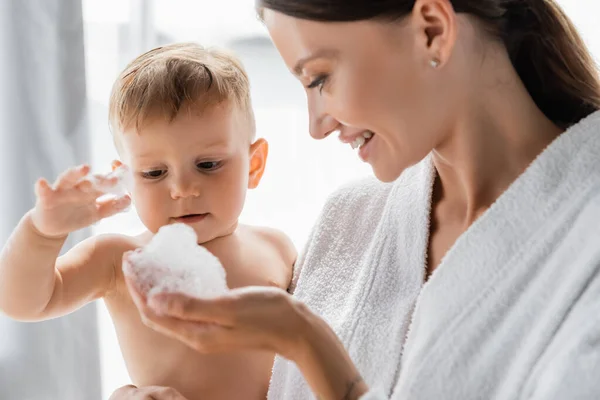Tout-petit garçon touchant mousse de bain dans la main de la mère heureuse en peignoir — Photo de stock