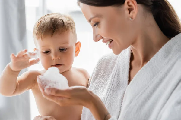 Tout-petit garçon touchant mousse de bain dans la main de la mère heureuse en peignoir — Photo de stock