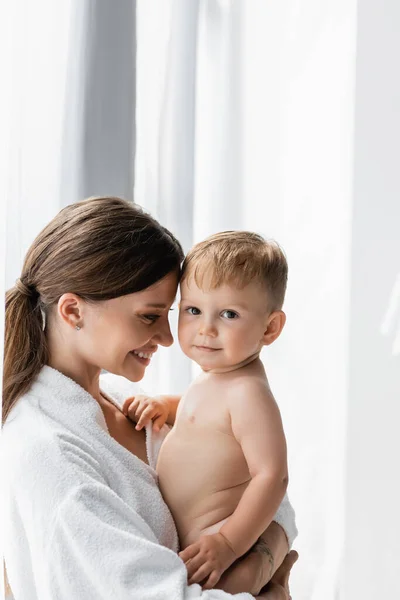 Smiling and tattooed mother in bathrobe holding in arms nude toddler son — Stock Photo