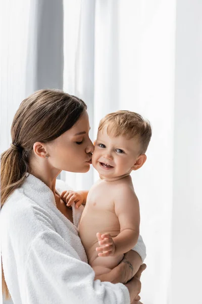 Tattooed mother in bathrobe holding in arms and kissing cheek of nude toddler son — Stock Photo