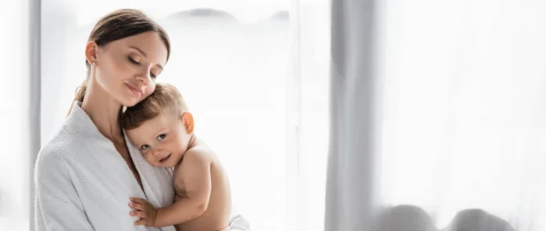 Pleased mother in bathrobe holding in arms nude toddler son, banner — Stock Photo