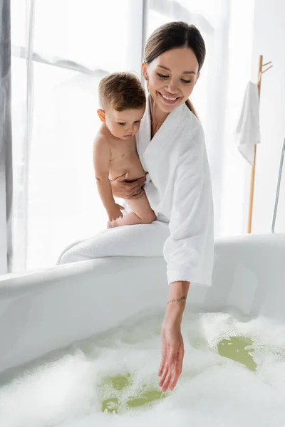 Pleased and tattooed mother in bathrobe holding in arms nude toddler son and reaching water in bathtub — Stock Photo