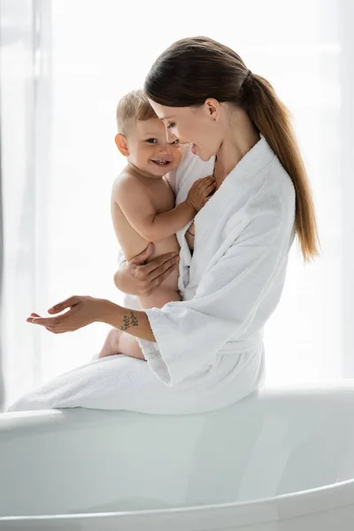 Pleased and tattooed mother in bathrobe holding in arms cheerful toddler son near bathtub — Stock Photo