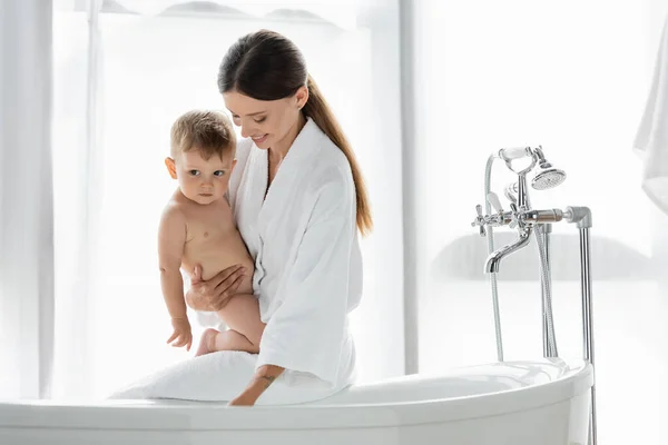 Pleased woman in bathrobe holding in arms toddler son near bathtub — Stock Photo