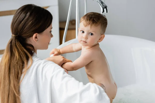 Мати в халаті дивиться на маленького хлопчика у ванній — стокове фото