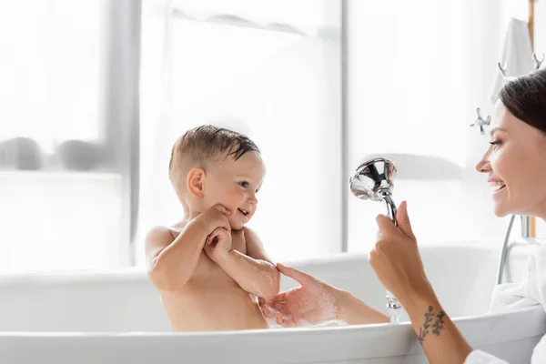 Alegre madre con tatuaje celebración ducha cabeza mientras bañando niño en bañera - foto de stock