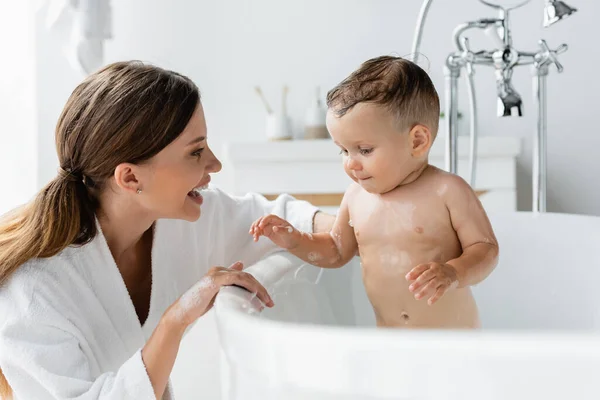 Alegre madre en albornoz bañándose mojado niño hijo en bañera - foto de stock
