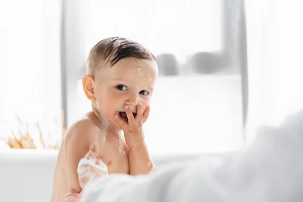 Blurred mother in bathrobe bathing wet and happy toddler son in bathtub — Stock Photo