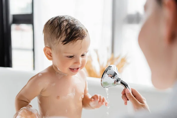 Madre borrosa sosteniendo la cabeza de la ducha y bañando a su hijo pequeño - foto de stock