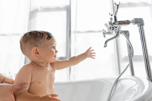 Tattooed mother holding toddler son reaching faucet while bathing in bathtub — Stock Photo