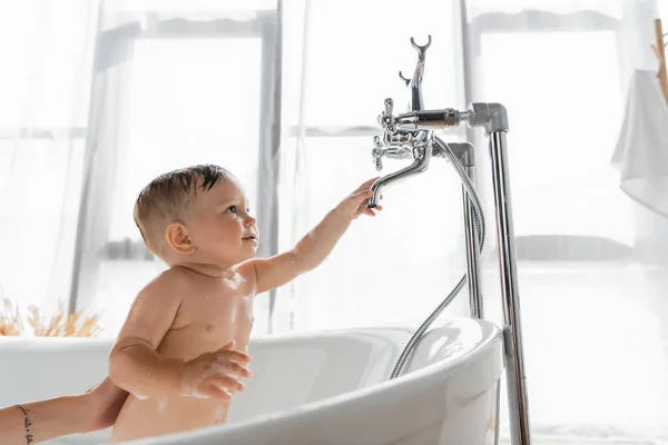 Tattooed mother holding toddler son reaching faucet while bathing in bathroom — Stock Photo
