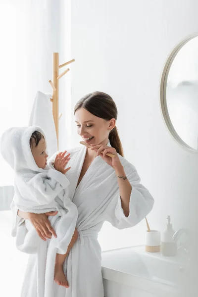 Happy mother brushing teeth and holding in arms toddler son in bathrobe — Stock Photo