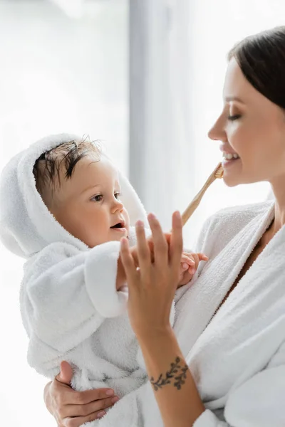 Toddler son in bathrobe helping happy mother brushing teeth — Stock Photo