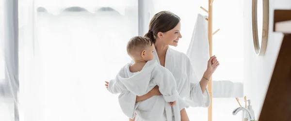 Seitenansicht der fröhlichen Mutter, die ihren kleinen Sohn im Arm hält und im Badezimmer auf den Spiegel schaut, Banner — Stockfoto