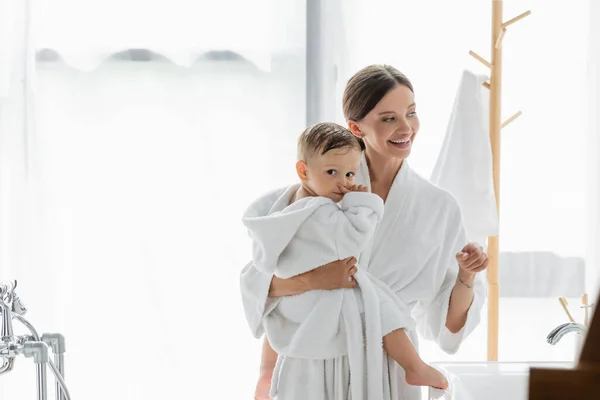 Cheerful mother holding in arms toddler son and looking away in bathroom — Stock Photo