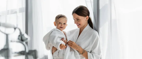 Cheerful mother looking at toddler son in bathrobe, banner — Stock Photo