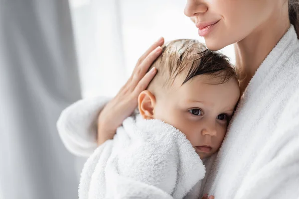 Pleased young mother holding in arms toddler son in bathrobe — Stock Photo