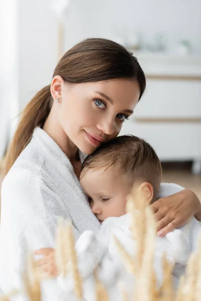 Madre joven abrazando a su hijo pequeño en albornoz - foto de stock