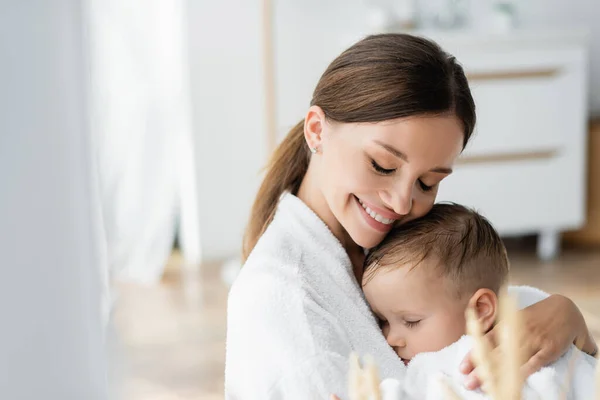 Sonriente joven madre con los ojos cerrados abrazando a su hijo pequeño en albornoz - foto de stock