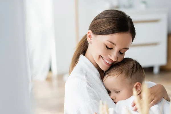 Souriant jeune mère avec les yeux fermés étreignant tout-petit fils en peignoir — Photo de stock