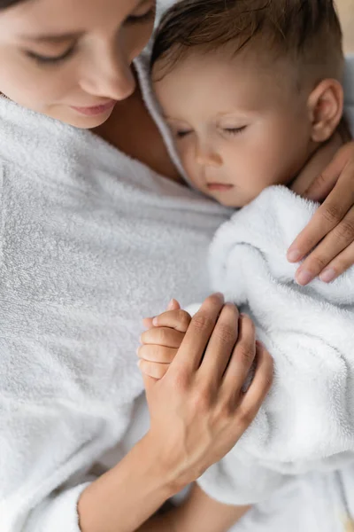 Madre borrosa y cariñosa abrazando a su hijo pequeño con los ojos cerrados en albornoz - foto de stock