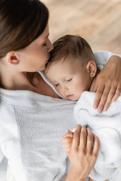 Caring mother kissing head of toddler son with closed eyes in bathrobe — Stock Photo