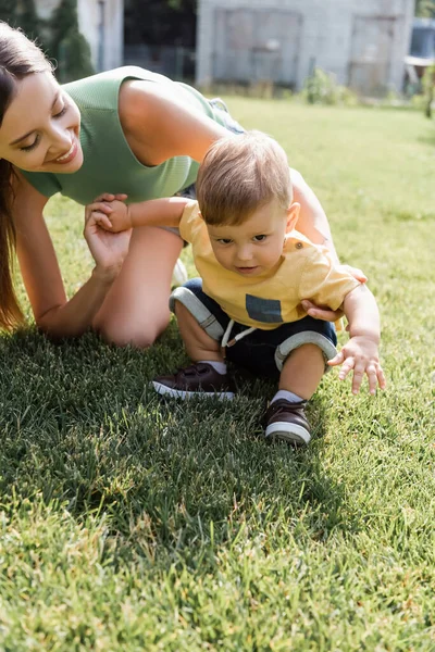 Feliz madre mirando a su hijo pequeño sentado en la hierba verde - foto de stock