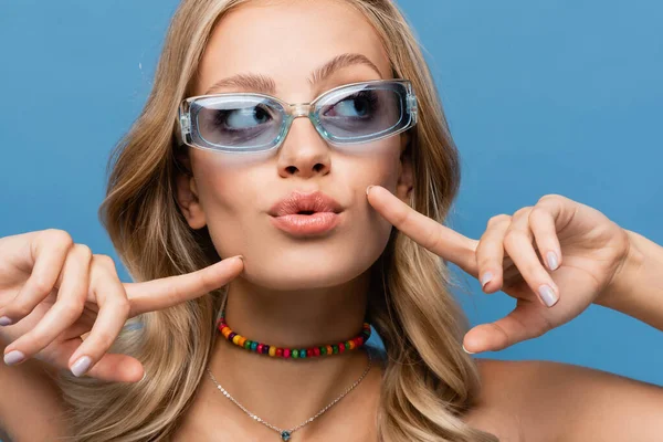 Mujer joven en gafas de sol de moda haciendo pucheros labios y apuntando a las mejillas aisladas en azul - foto de stock