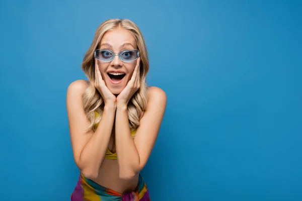 Здивована молода жінка в модних сонцезахисних окулярах дивиться на камеру ізольовано на синьому — стокове фото