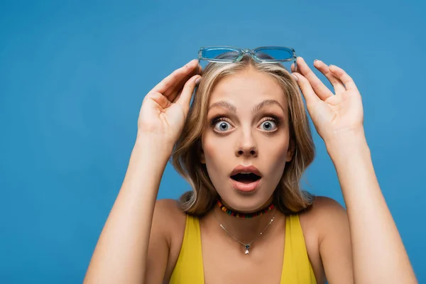 Scioccato giovane donna regolazione occhiali da sole alla moda e guardando la fotocamera isolata su blu — Foto stock