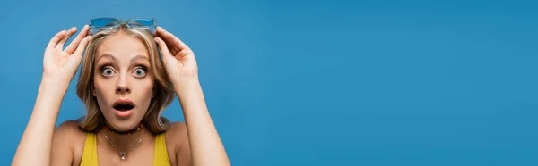 Chocado jovem mulher ajustando óculos de sol na moda e olhando para a câmera isolada em azul, banner — Fotografia de Stock