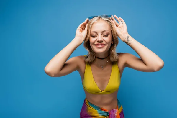 Heureux jeune femme en haut de bikini jaune réglage des lunettes de soleil à la mode isolé sur bleu — Photo de stock