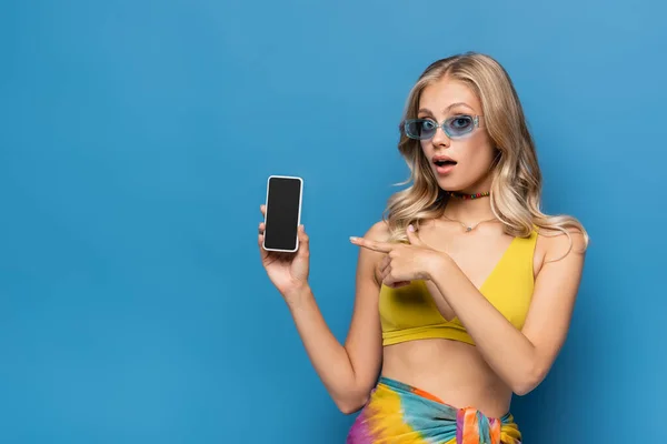 Surpresa jovem mulher em biquíni amarelo top apontando para smartphone com tela em branco isolado em azul — Fotografia de Stock