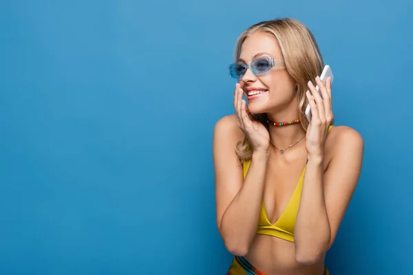 Mujer joven feliz en bikini amarillo superior hablando en el teléfono inteligente aislado en azul - foto de stock