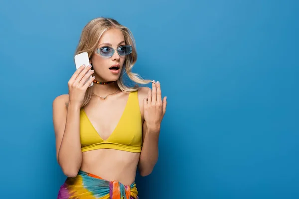 Shocked young woman in yellow bikini top talking on smartphone isolated on blue — Stock Photo