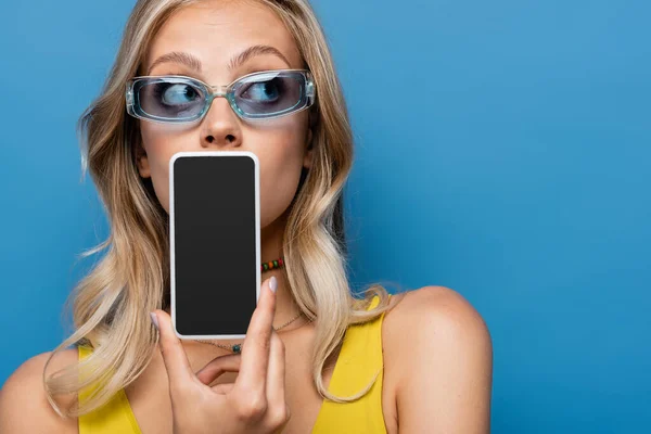 Молодая блондинка в солнечных очках, прикрывающая рот, держа смартфон с пустым экраном, изолированным на синем — стоковое фото