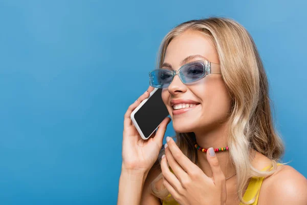 Joyful young woman with beaded necklace talking on smartphone isolated on blue — Stock Photo