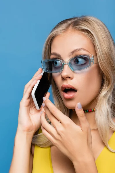 Шокована молода жінка в сонцезахисних окулярах говорить на смартфоні ізольовано на синьому — стокове фото