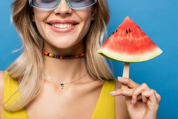 Abgeschnittene Ansicht einer glücklichen jungen Frau mit Sonnenbrille, die Eis am Stiel mit Wassermelone auf blauem Grund hält — Stockfoto