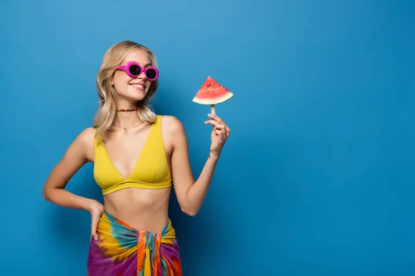 Glückliche junge Frau, die mit der Hand auf der Hüfte steht und Eis am Stiel mit Wassermelone auf blau hält — Stockfoto