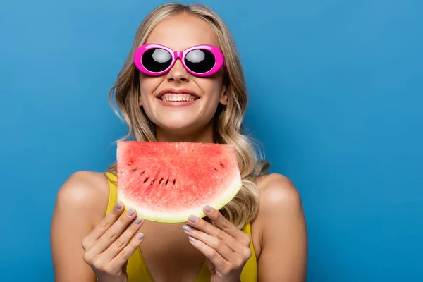 Радісна молода жінка в рожевих сонцезахисних окулярах тримає шматочок кавуна ізольовано на синьому — стокове фото