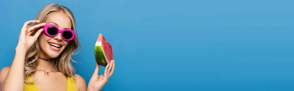 Joyful young woman adjusting pink sunglasses and holding slice of watermelon isolated on blue, banner — Stock Photo