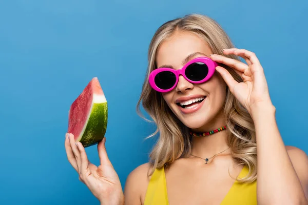 Радісна молода жінка регулює рожеві сонцезахисні окуляри і тримає шматочок кавуна ізольовано на синьому — стокове фото