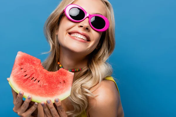 Весела молода жінка в рожевих сонцезахисних окулярах тримає солодкий кавун ізольовано на синьому — стокове фото