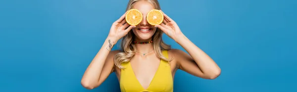 Cheerful young woman in yellow bikini top covering eyes with orange halves isolated on blue, banner — Stock Photo