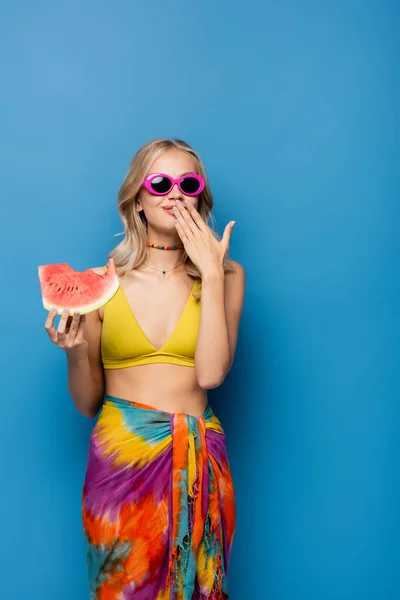 Sonriente joven en gafas de sol de color rosa cubriendo la boca mientras come sandía fresca en azul - foto de stock