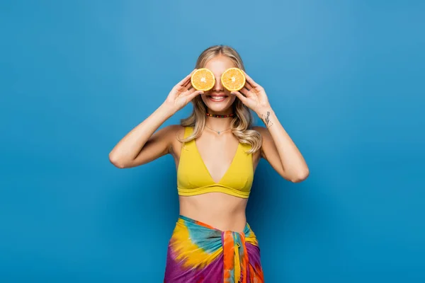 Cheerful young woman in yellow bikini top covering eyes with orange halves on blue — Stock Photo
