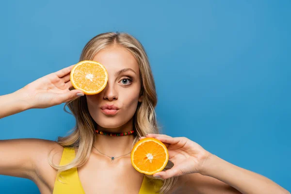 Rubia joven mujer haciendo pucheros labios y cubriendo el ojo con naranja medio aislado en azul - foto de stock
